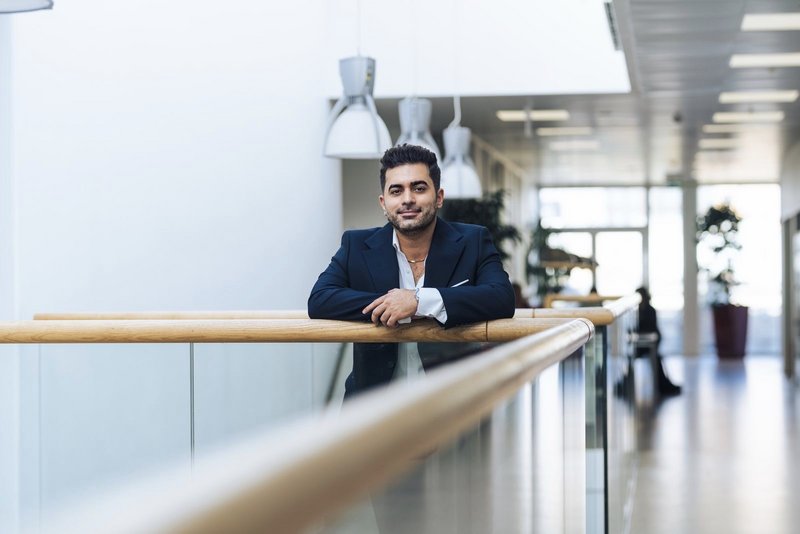 Behzad Bozorgtabar leaning on a wooden railing in a bright corridor at Aarhus University.